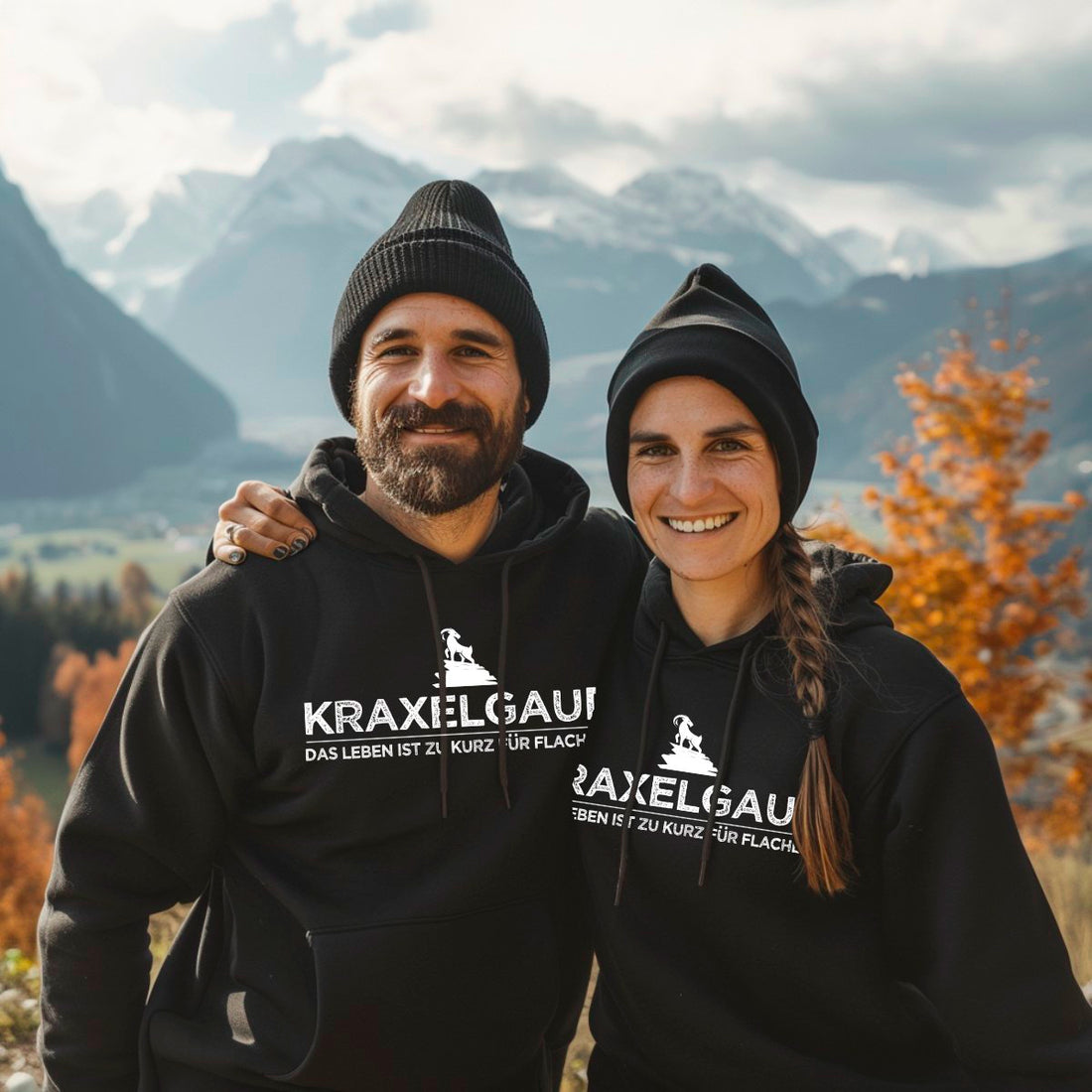 Paar in schwarzen Kraxelgaudi Hoodies mit Mützen vor herbstlichem Bergpanorama