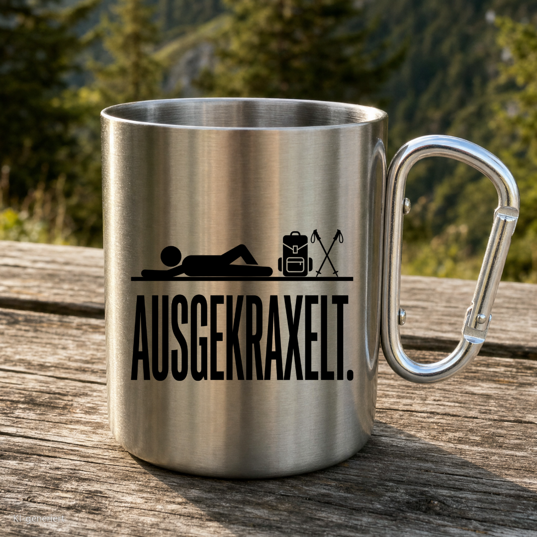 Karabinertasse von Kraxelgaudi mit Motiv Ausgekraxelt steht auf Holz vor Bergkulisse
