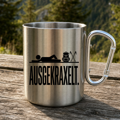 Karabinertasse von Kraxelgaudi mit Motiv Ausgekraxelt steht auf Holz vor Bergkulisse