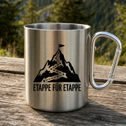 Karabinertasse von Kraxelgaudi mit Motiv Etappe für Etappe steht auf Holz vor Bergkulisse