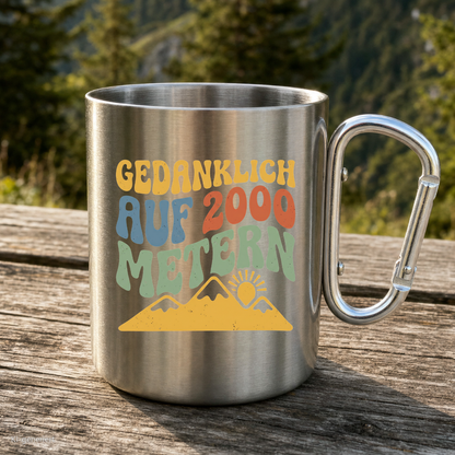 Karabinertasse von Kraxelgaudi mit Spruch Gedanklich auf 2000 Metern steht auf Holz vor Bergkulisse