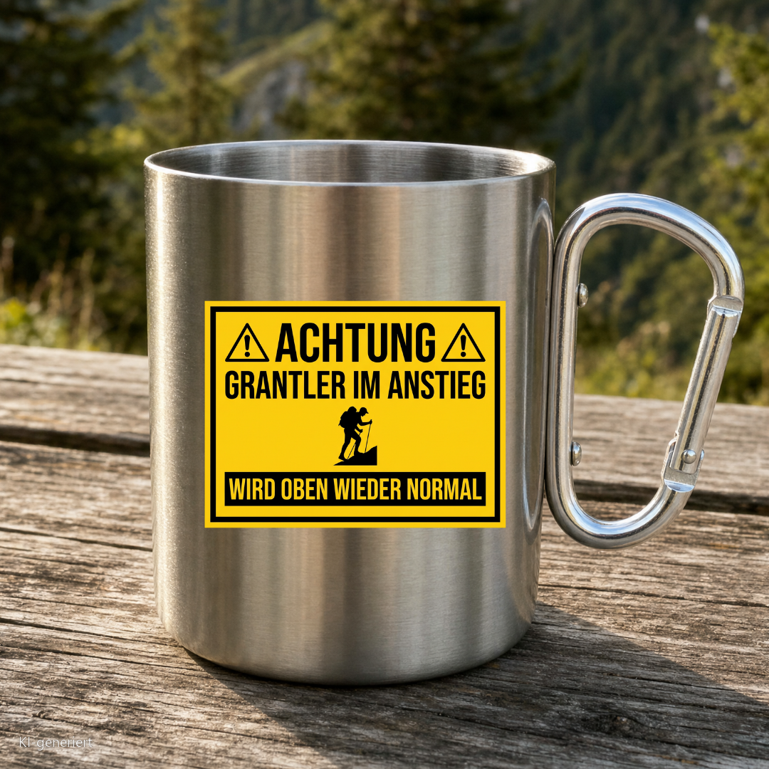 Karabinertasse von Kraxelgaudi mit Warnschild Grantler im Anstieg steht auf Holz vor Bergkulisse