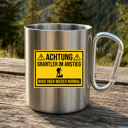 Karabinertasse von Kraxelgaudi mit Warnschild Grantler im Anstieg steht auf Holz vor Bergkulisse