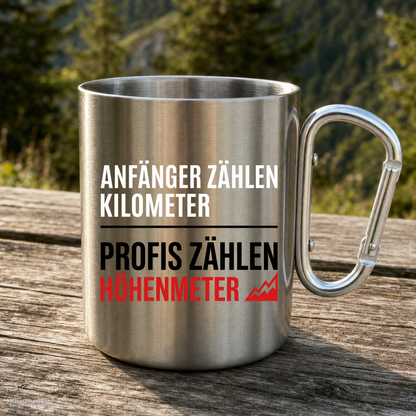 Karabinertasse von Kraxelgaudi mit Spruch Profis zählen Höhenmeter steht auf Holz vor Bergkulisse