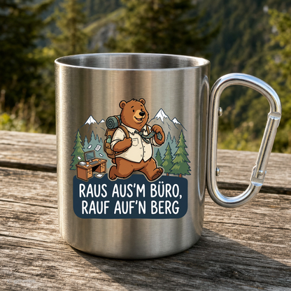 Karabinertasse von Kraxelgaudi mit Bär und Spruch Raus ausm Büro rauf aufn Berg steht auf Holz vor Bergkulisse