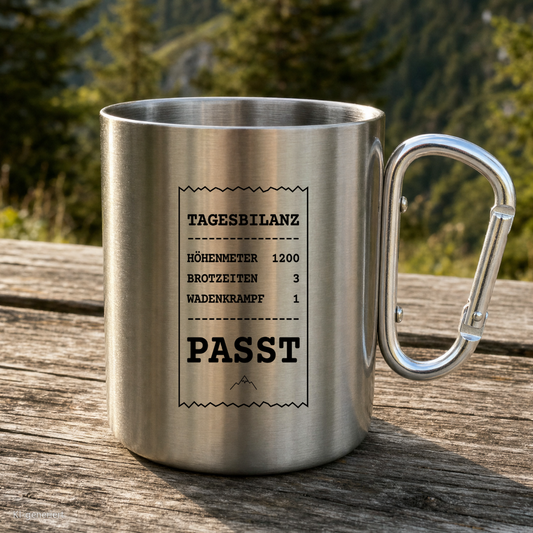 Karabinertasse von Kraxelgaudi mit Tagesbilanz Motiv steht auf Holz vor Bergkulisse